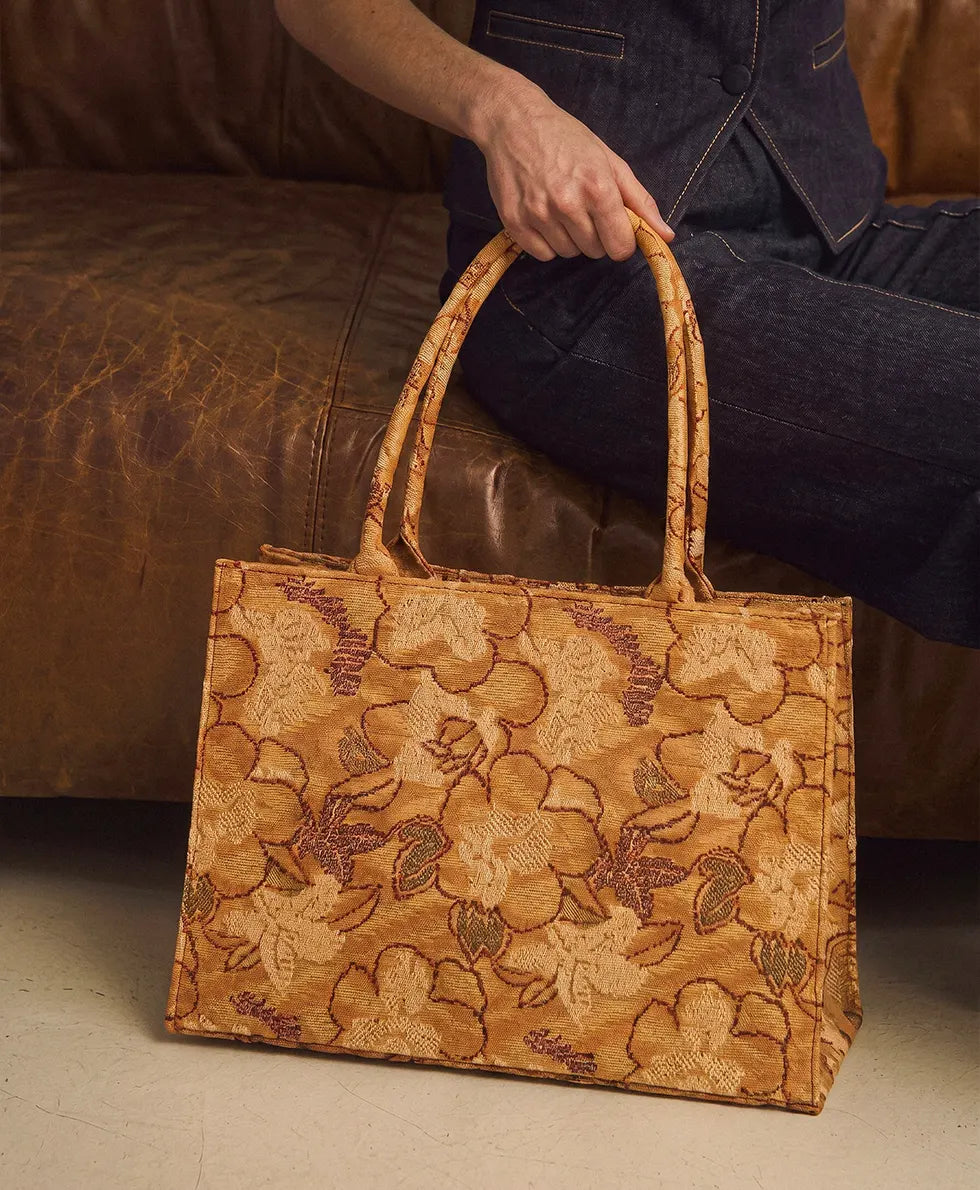 A women sitting on a brown leather couch holds a large tan woven tote bag with floral embroidery and dual top handles.