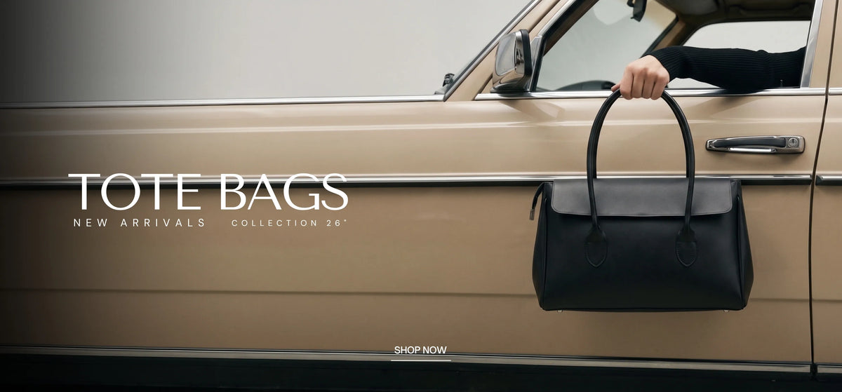 A black leather tote bag with a gray flap and double top handles is held by a person's hand next to a beige vintage car.