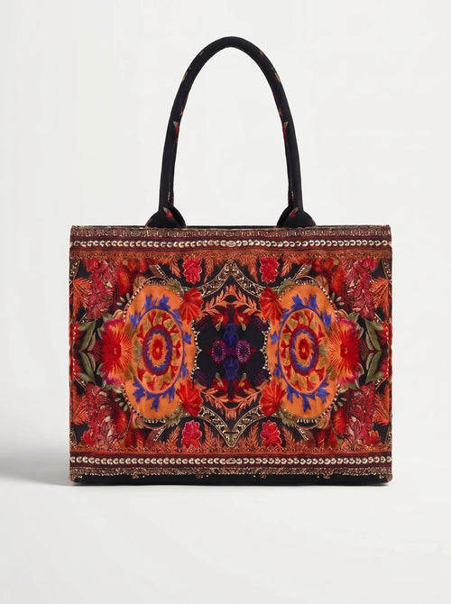 A structured tote bag with a vibrant, symmetrical multicolor floral and geometric pattern, featuring black shoulder straps and a beaded border, set against a plain light gray background.